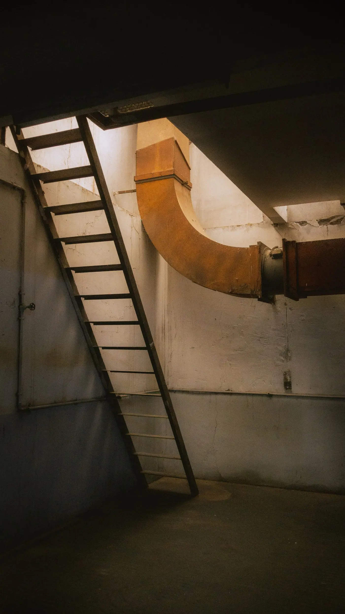 Large ductwork elbow and a step ladder in a dim basement mechanical space