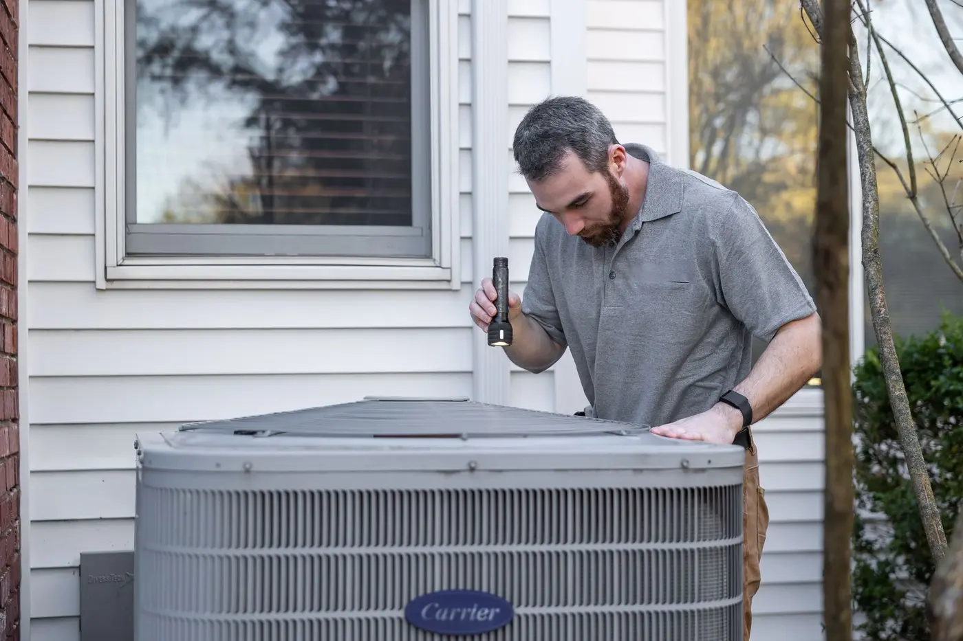 Technician checking an outdoor heat pump unit with a flashlight beside a house