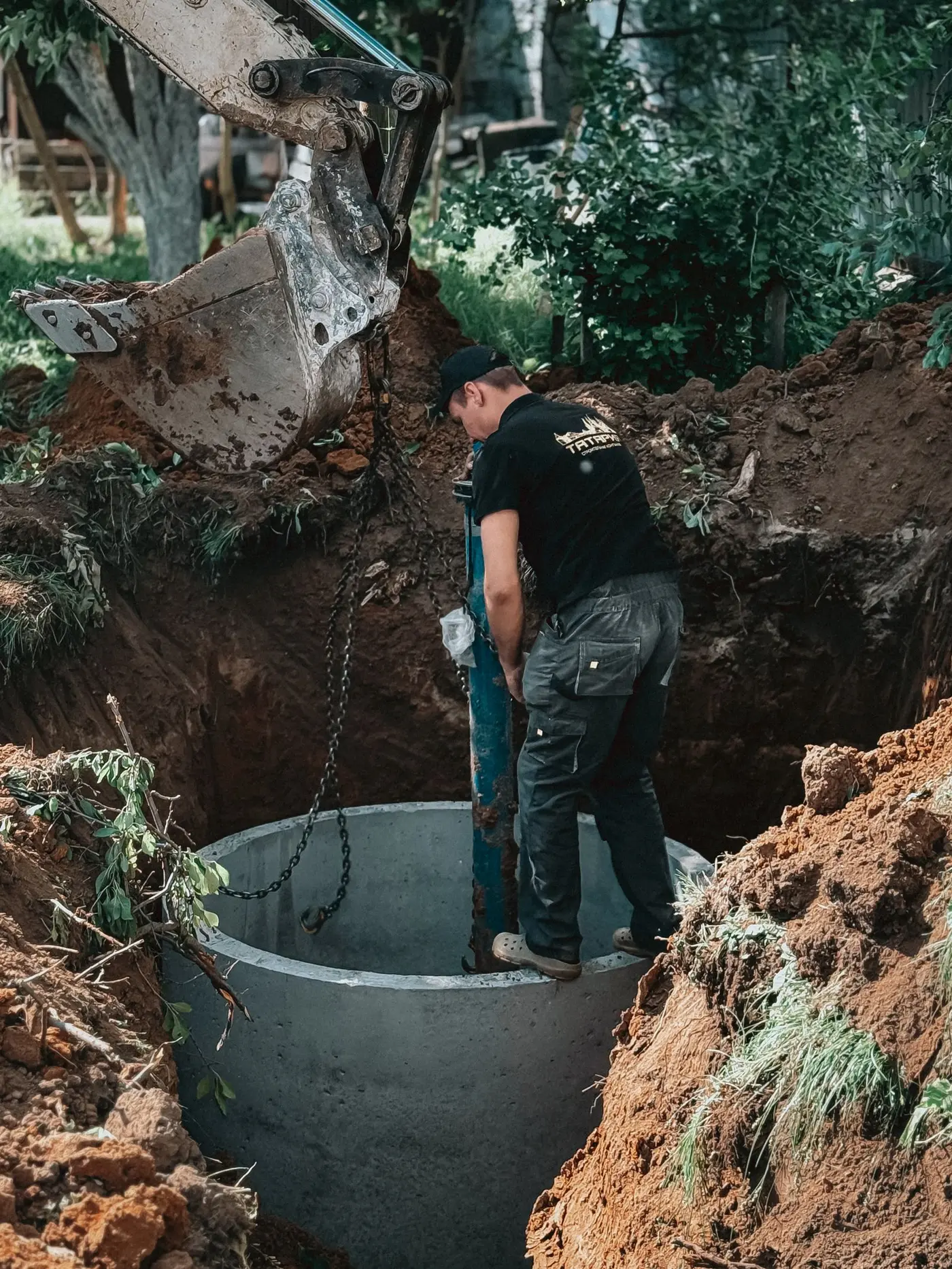 Excavator lowering a concrete service ring into a dug pit while a worker guides it